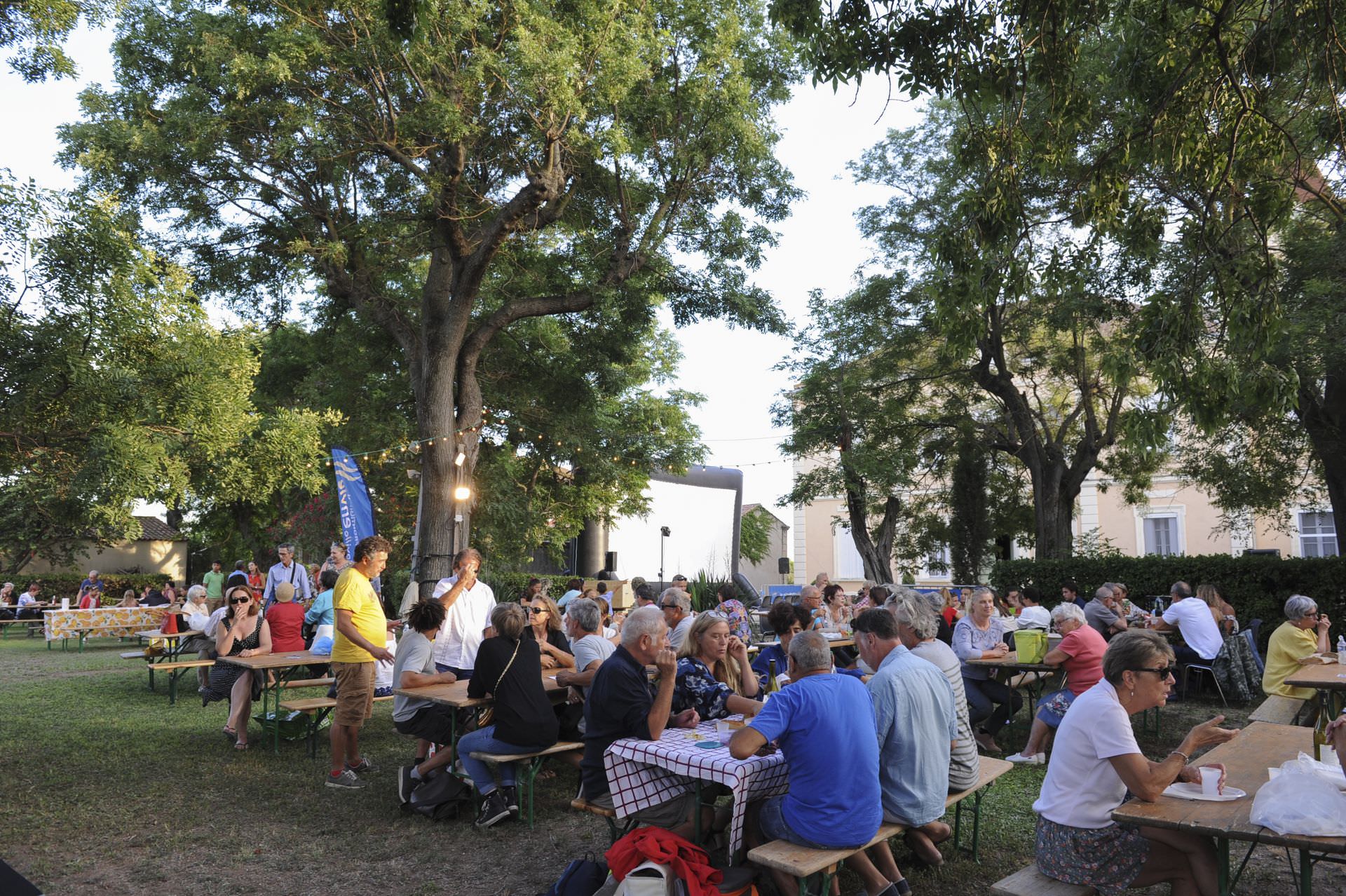 Personnes attablées lors du Festival Emmuscades sour les arbres du Château Lapeyrade à Frontignan, toile de cinéma en arrière-plan
