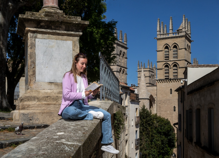 Jeune fille assise sur l'e mur de l'esplanade de la Canourgue lisant un livre. Vur sur la Cathédrale Saint Pierre de Montpellier