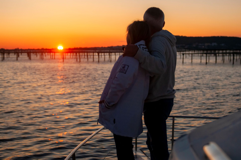 Un couple sur le bateau Mansathau, admire le coucher de soleil sur l'étang de Thau. Sète et les tables des conchyliculteurs se détachent grâce aux couleurs orangées du soleil couchant.